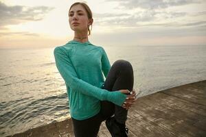 Portrait of young woman in bright sportswear, trains at the seaside, makes morning stretching exercises before running. Health care concept. photo