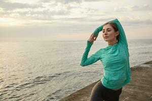 Young sporty lady in bright sportswear, trains at the seaside, makes morning stretching exercises after running. Health care concept. photo