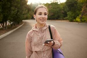 Young positive woman walking after yoga at the park and chatting wit friend, hols the smartphone in hand, listening music in headphones, looks away and smiling. photo