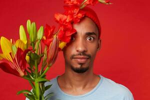 joven conmocionado atractivo chico en rojo sombrero y azul camiseta, sostiene un ramo de flores en su manos y cubierto parte de cara con flores, mira a el cámara con amplio abierto ojos, soportes terminado rojo fondo foto