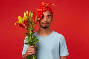 joven sorprendido atractivo hombre en rojo sombrero y azul camiseta, sostiene un ramo de flores en su manos y cubierto parte de cara con flores, miradas a el cámara con amplio abierto ojos, soportes terminado rojo fondo foto