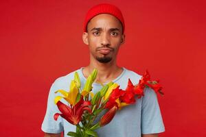 retrato de joven infeliz atractivo chico en rojo sombrero y azul camiseta, sostiene un ramo de flores en su manos, mira a el cámara con triste expresión, soportes terminado rojo fondo foto