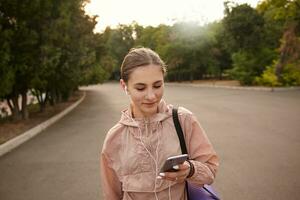 retrato de joven alegre mujer caminando después yoga a el parque y chateando ingenio amigo, mira a el teléfono inteligente en mano, escuchando música en auriculares. foto