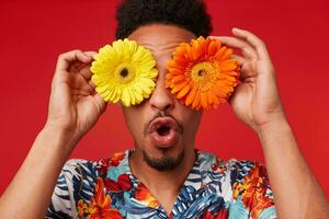 Close up surprised young African American guy, wears in Hawaiian shirt, looks at the camera trough flowers with happy expression, stands over red background. photo