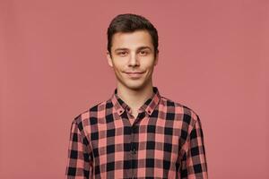Portrait of young attractive man in checkered shirt, calmly looks at the camera, stands over pink background and smiles. photo