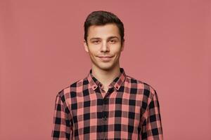 retrato de joven atractivo hombre en a cuadros camisa, tranquilamente mira a el cámara, soportes terminado rosado antecedentes y sonrisas foto