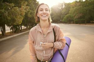 retrato de joven bonito niña caminando después yoga a el parque, escuchando música en auriculares, siente genial y sonriente, disfrutar el frio día. foto