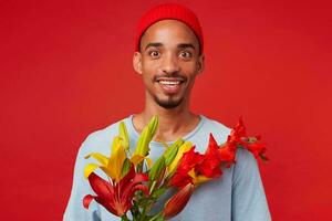 retrato de joven alegre atractivo chico en rojo sombrero y azul camiseta, sostiene un ramo de flores en su manos, mira a el cámara con contento expresión y sonriente, soportes terminado rojo fondo foto