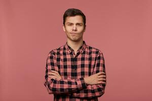 Young handsome discontent guy wears in checkered shirt, frowningn and looks at the camera with crossed arms, isolated over pink background. photo