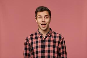 Portrait of young attractive happy amazed man in checkered shirt, looks at the camera with surprised expression, stands over pink background with wide open mouth. photo