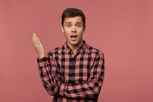 Young indigant guy wears in checkered shirt, looks at the camera with surprised expression, looks shocked, isolated over pink background. photo