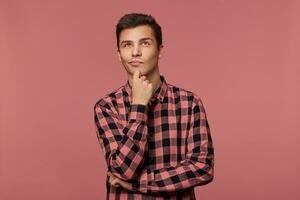 Young handsome pensive guy wears in checkered shirt, looks up with and touches chin, thinks about something, isolated over pink background. photo