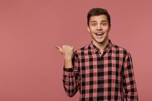 joven atractivo alegre hombre usa en a cuadros camisa, mira a el cámara con contento expresión, quiere a giro tu atención a Copiar espacio a el izquierda lado, soportes terminado rosado antecedentes. foto