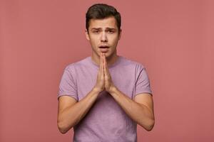 joven atractivo chico usa en blanco camiseta, hacer un desear, esperanzas para bueno suerte en exámenes y muestra orado gesto, soportes terminado rosado antecedentes. foto