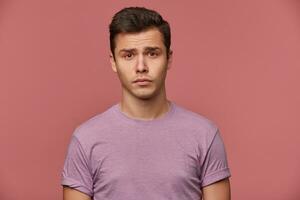 Close up of young offended attractive boy in blank t-shirt, stands over pink background and looks sad and unhappy. photo