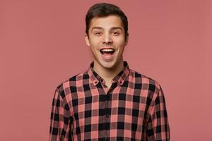 Portrait of young attractive surprised man in checkered shirt, looks at the camera with wide open mouth, stands over pink background. photo