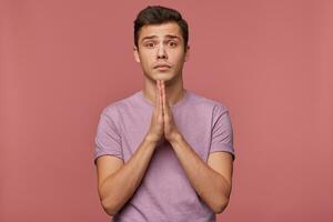 joven atractivo chico usa en blanco camiseta, hacer un desear, esperanzas para bueno suerte en exámenes y muestra orado gesto, pregunta no a dar él un malo marca, soportes terminado rosado antecedentes. foto