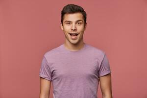 retrato de hermoso alegre chico usa en blanco camiseta, mira a el cámara con contento asombrado expresión, soportes terminado rosado antecedentes. foto