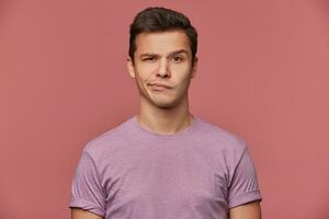 Portrait of handsome frowning young man wears in blank t-shirt, looks at the camera with with a grin and doubts, stands over pink background. photo