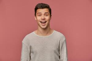 Portrait of cheerful handsome young guy wears in blank long sleeve, looks at the camera with happy expression, stands over pink background. photo