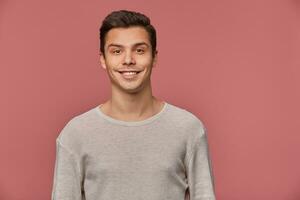 Photo of smiling handsome young guy wears in blank long sleeve, looks cheerful and glad, looks at the camera with happy expression, stands over pink background and smiling.