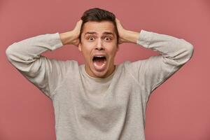 Close up of young scared man in blank long sleeve, stands over pink background with closed eyes and screaming, looks afraid and unhappy, touches head. photo