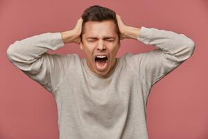Close up of young attractive man in blank longs leeve, stands over pink background with closed eyes and screaming, looks angry and unhappy, touches head. photo