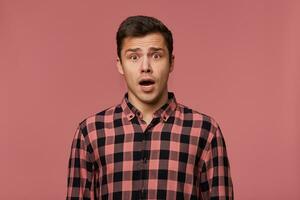 Young handsome amazed man in checkered shirt, looks at the camera with with wide open mouth in surprised expression, isolated over pink background. photo