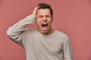 Photo of young attractive guy in blank long sleeve, stands over pink background with closed eyes and screaming, looks angry and unhappy.