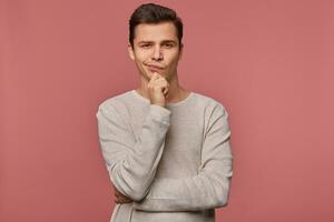 Young handsome thinking guy wears in checkered shirt, looks at the camera and touches chin, thinks about something, isolated over pink background. photo