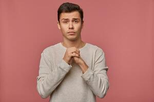 Young attractive guy wears in blank long sleeve, make a wish, hopes for good luck on exams and crossed palms, asks not to give him a bad mark, stands over pink background. photo