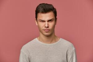 Close up of young handsome strict guy wears in basic long sleeve, looks at the camera with angry expression, isolated over pink background. photo