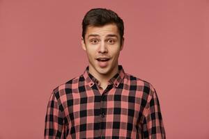 Young handsome wondered guy wears in checkered shirt, looks at the camera with with wide open mouth in surprised expression, isolated over pink background. photo