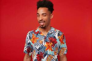 Young positive African American man wears in Hawaiian shirt, looks at the camera and winks, stands over red background. photo