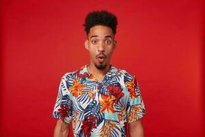 Portrait of shocked young African American guy, wears in Hawaiian shirt, looks at the camera with surprised expression with wide open mouth and eyes, stands over red background. photo