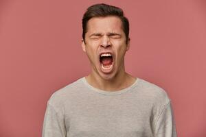 Close up of young evil attractive guy wears in blank long sleeve, stands over pink background with closed eyes and screaming, looks angry and unhappy. photo