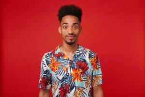 joven africano americano hombre usa en hawaiano camisa, mira a el cámara y calma sonriente, soportes terminado rojo antecedentes. foto