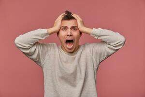 Young scared man in blank long sleeve, holds head and looks at the camera with shocked expression, stands over pink background and screaming, looks afraid. photo