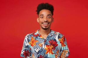 retrato de alegre joven africano americano chico, usa en hawaiano camisa, mira a el cámara con contento expresión, soportes terminado rojo antecedentes y en general sonrisas foto