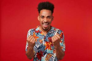 Photo of young joyful African American man, wears in Hawaiian shirt, looks happy and glad, clenches fists and rejoices victory, stands over red background.