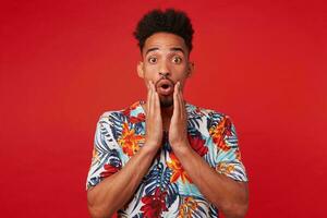 Young shocked African American guy wears in Hawaiian shirt, looks at the camera with surprised expression, stands over red background and touches cheeks. photo