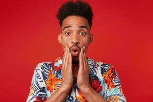 Close up of young shocked African American guy wears in Hawaiian shirt, looks at the camera with surprised expression, with wide open mouth, stands over red background and touches cheeks. photo