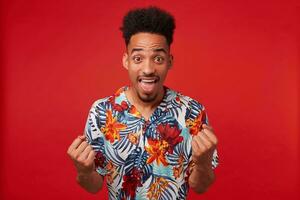 Photo of young happy African American man, wears in Hawaiian shirt, looks at the camera with wide open mouth, clenches fists and rejoices victory, stands over red background.