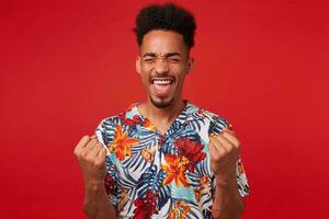 joven africano americano chico, usa en hawaiano camisa, mira contento y contento, soportes terminado rojo antecedentes y aprieta puños y se regocija victoria. foto