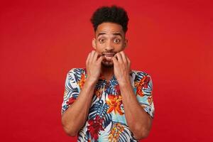 Young scared African American guy wears in Hawaiian shirt, looking at the camera and bites nails, stands over red background. photo