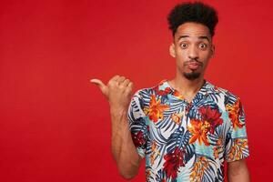 Portrait of young African American man in Hawaiian shirt, looks amazed and happy, stands over red background, points to the left at copyspace. photo