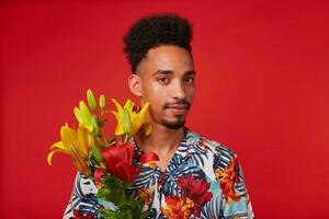 Close up of calm young African American man, wears in Hawaiian shirt, looks at the camera, holds yellow and red flowers, stands over red background. photo