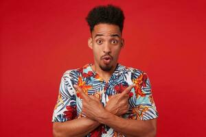 Portrait if young shocked African American man in Hawaiian shirt, looks at the camera with surprised expression, stands over red background, points in different directions. photo