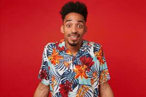 retrato de alegre joven africano americano chico, usa en hawaiano camisa, mira a el cámara con contento expresión, soportes terminado rojo antecedentes y en general sonrisas foto