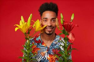 Young African American guy, wears in Hawaiian shirt, looks at the camera with happy expression, stands over red background with yellow and red flowers and smiles. photo